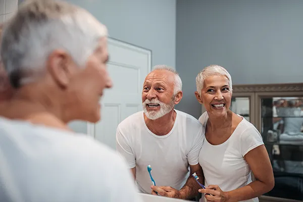 Elderly white couple smiling and brushing their teeth in the bathroom mirror