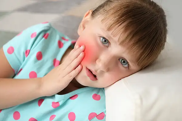 Little girl lying down and holding her red, swollen cheek from a severe toothache, indicating the need for emergency dental treatment for kids.