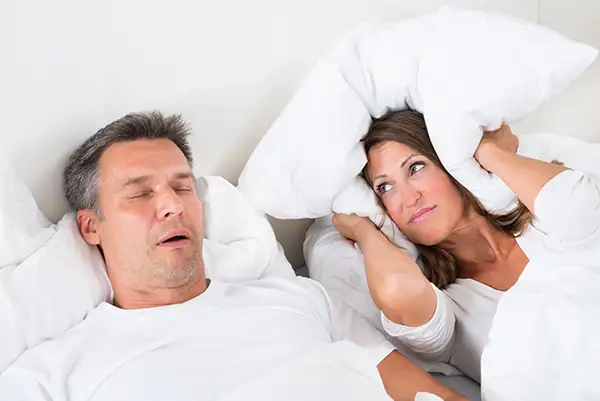 A husband snoring very loudly while his wife covers her ears with a pillow in frustration, illustrating the impact of sleep apnea on daily life and relationships.